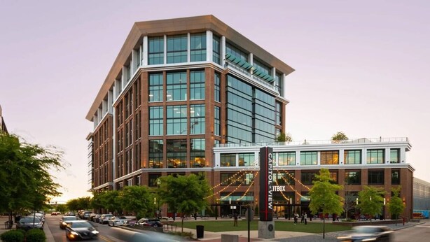 A modern building with a glass facade, a prominent entrance with a sign, and a parking lot with cars.