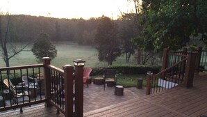 Una terraza de madera con mobiliario exterior, barandilla y vista a un lago y un bosque.