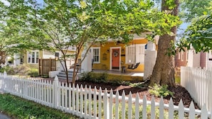 A yellow house with a white picket fence, a porch, and a tree in front. - Carolina Cottage at Carolina Pl (Wilmington)
