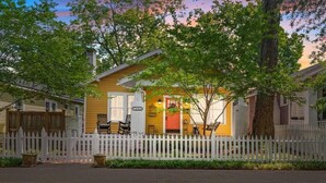 A yellow house with a white picket fence, a porch with chairs, and a small tree in front.