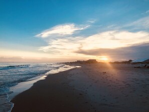 Ein Strand mit Sonnenuntergang, Wellen und einigen Gebäuden in der Ferne.