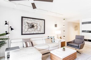 A modern living room with a white sofa, a gray armchair, and a coffee table. There is a large abstract painting on the wall, a floor lamp, and a small side table with a plant.