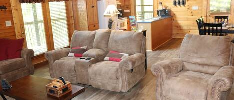A cozy log cabin interior with a living room featuring two sofas and a coffee table, a kitchen area with a refrigerator and microwave, and a dining area with a table and chairs.