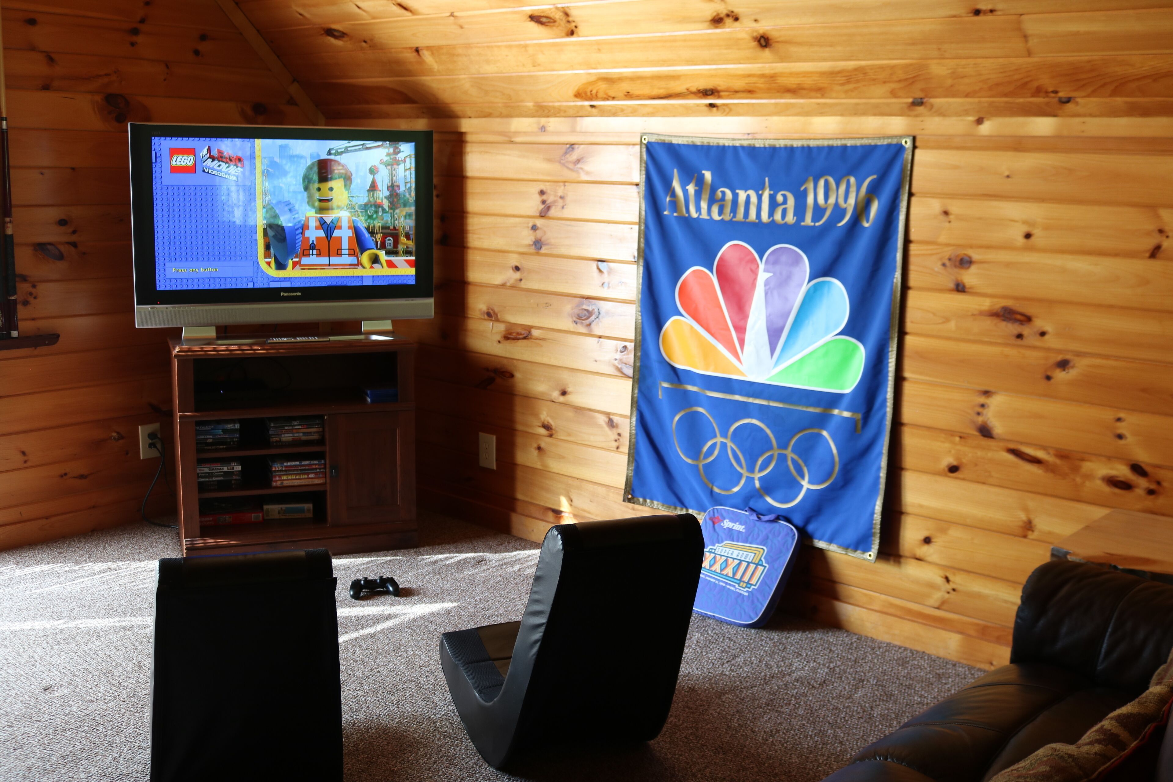 A room with a television displaying a video game, a banner with the NBC logo, and a wooden wall.