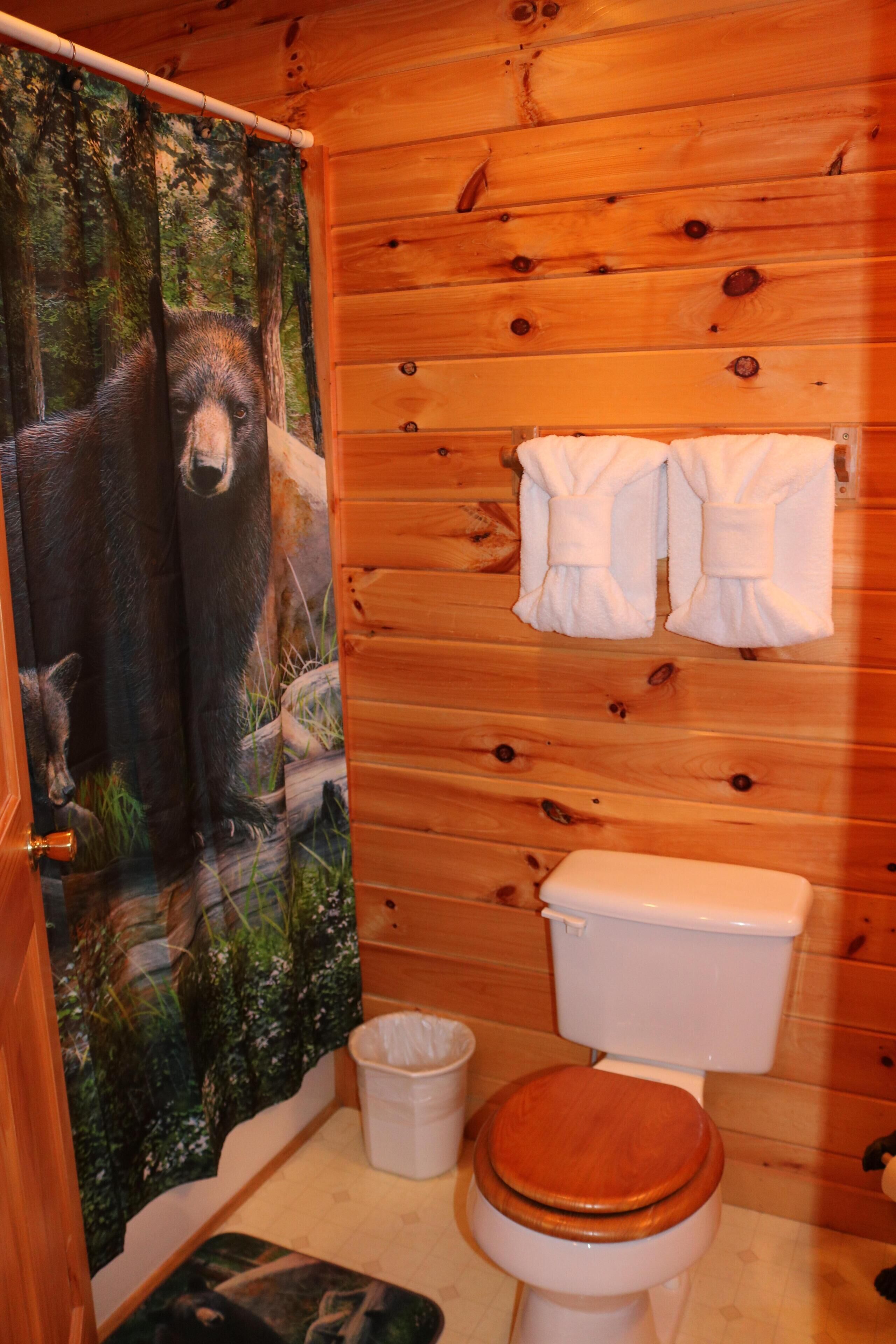 Une salle de bain avec un intérieur en bois, des toilettes blanches, un rideau de douche orné d’un motif d’ours et une paire de serviettes blanches accrochées au mur.