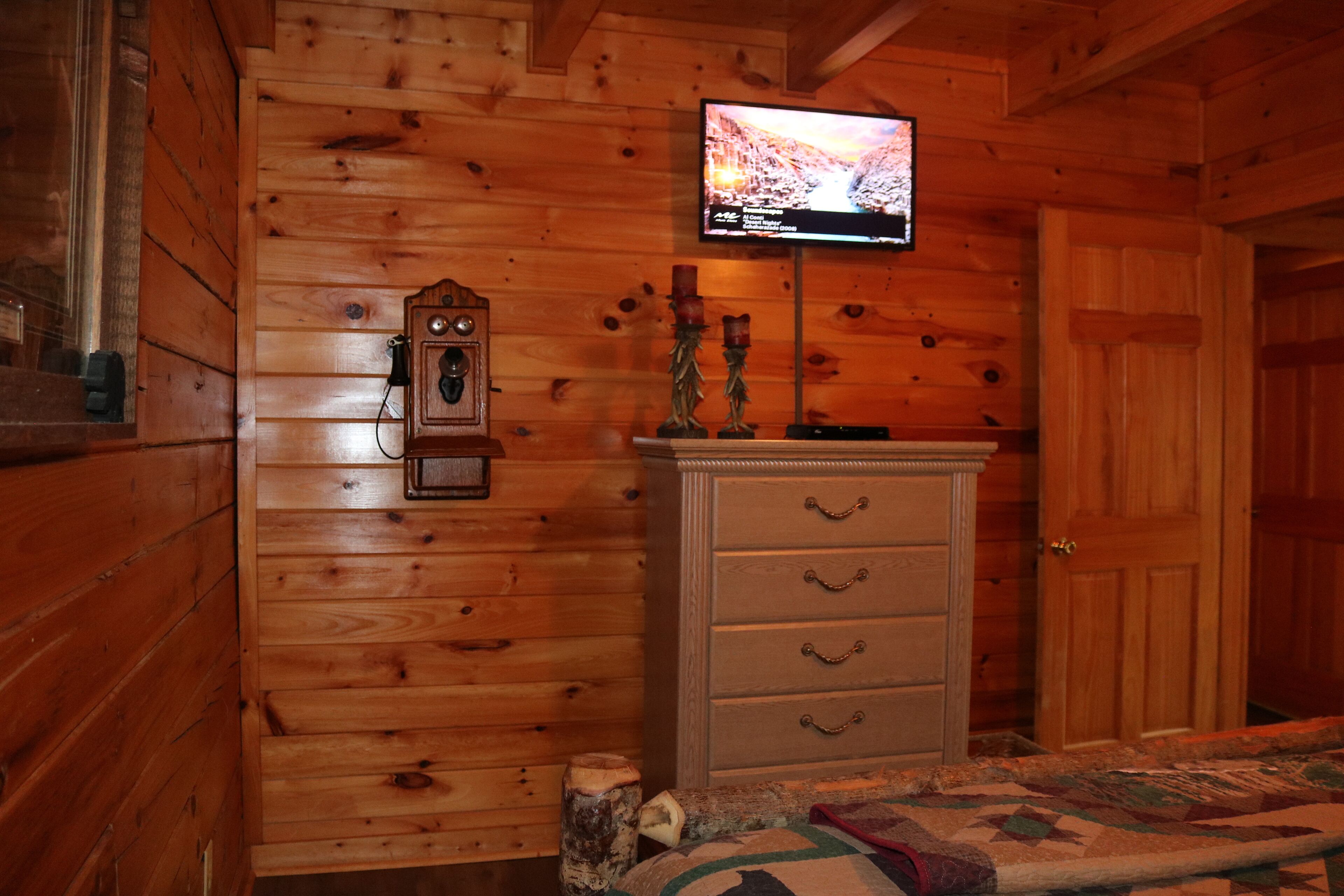 A wooden cabin room with a television mounted on the wall, a dresser with drawers, and a quilt on the bed.