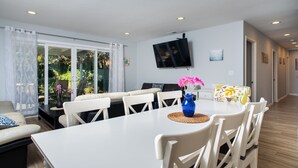 Un comedor con una mesa blanca, sillas blancas, un jarrón azul con flores rosadas y un televisor montado en la pared.