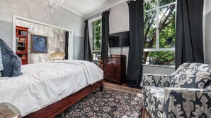 Een slaapkamer met een groot bed, een houten nachtkastje, een televisie, een boekenkast en een raam met gordijnen.
