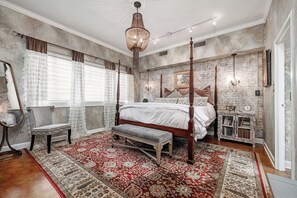 Un dormitorio con una cama con dosel, un banco, una mesita de noche y un amplio ventanal con cortinas.