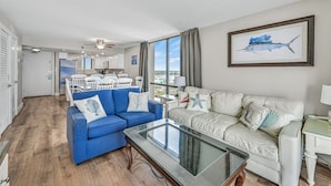 A modern living room with a blue sofa, a white sofa, and a glass coffee table. There is a kitchen in the background with white cabinets and a dining area.
