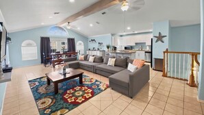 A spacious living area with a large gray sectional sofa, a coffee table, and a colorful rug. There is a kitchen in the background with white cabinets and a starfish decoration on the wall. - 2300 sq ft Beach close, spacious 5 star townhome, garage and parking! (South Padre Island)