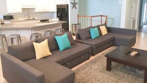 A modern living room with a gray sectional sofa, a coffee table, and a kitchen area with white cabinets and a black refrigerator. - 2300 sq ft Beach close, spacious 5 star townhome, garage and parking! (South Padre Island)