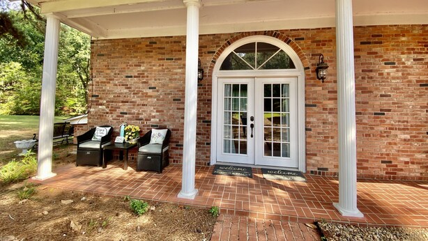 A covered patio with a brick wall, a white door with glass panels, and outdoor seating with cushions. - Your Creekside Villa Experience: Relax and Refresh. Brookhill Ranch Area!  (Hot Springs Village)