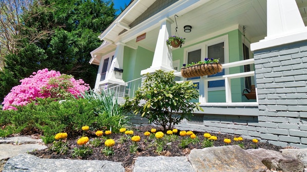 A house with a porch, a hanging flower basket, and a garden with yellow flowers and a pink bush.