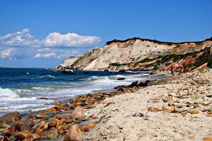 Une plage rocheuse où les vagues se brisent sur le rivage, une falaise couverte de végétation et un ciel dégagé.