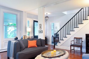 Amplia sala de estar con un sofá gris, una silla de madera y una bandeja con una vela. Una escalera con barandales negros conduce a otra habitación.
