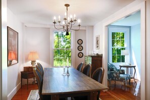 Comedor con mesa de madera, sillas, candelabro y ventanas amplias con follaje en el exterior.