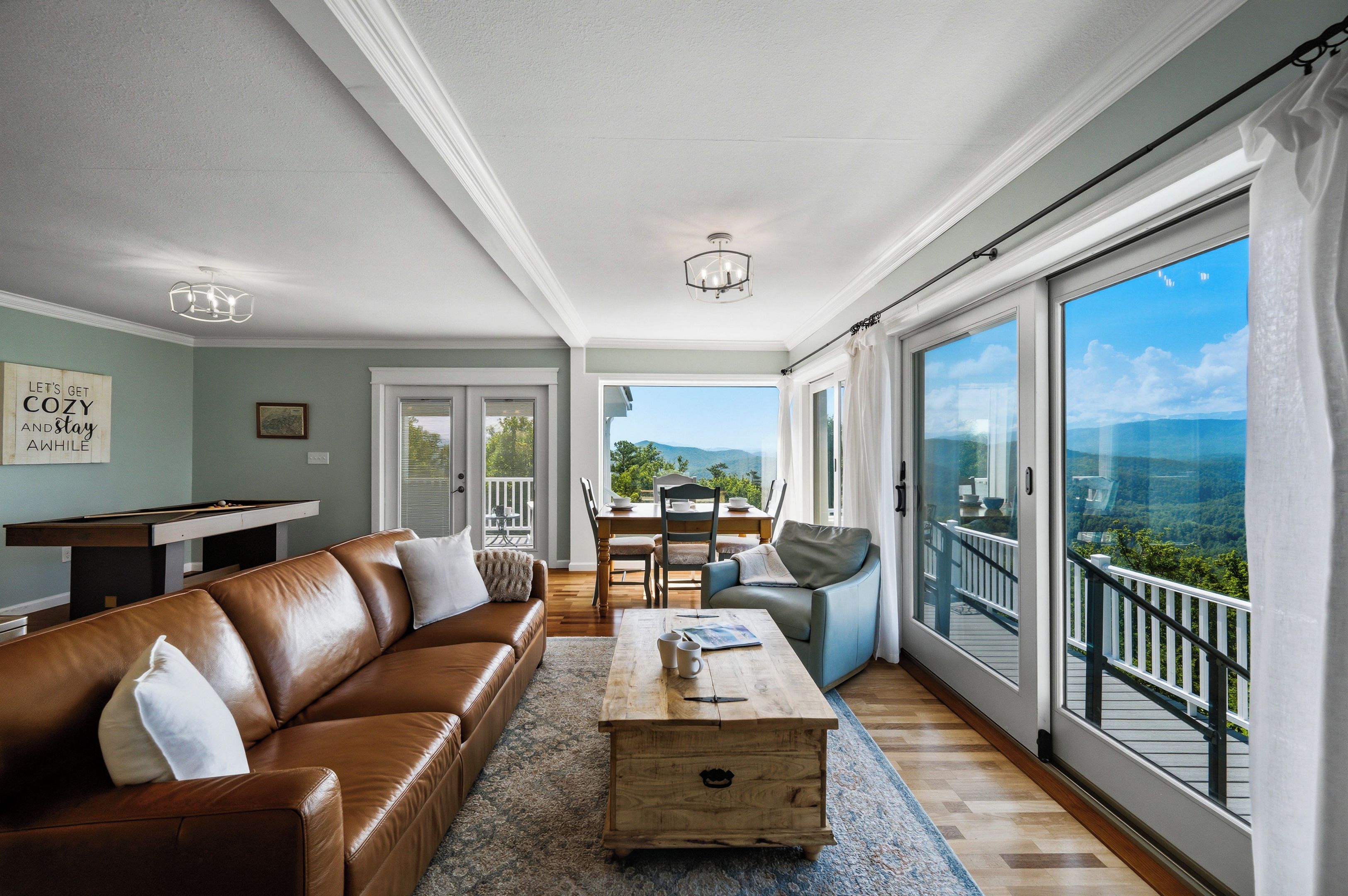 A spacious living room with a leather sofa, a wooden coffee table, and a view of a balcony with a scenic landscape.