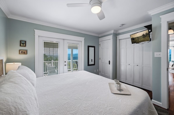 A bedroom with a large bed, a ceiling fan, a door leading to a balcony, and a television mounted on the wall.