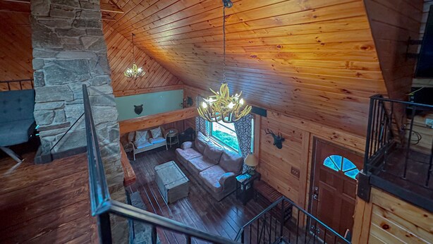 Ein gemütliches Blockhaus-Interieur mit Steinwand, Holzbalkendecke und Kamin. Eine Treppe führt zu einer oberen Ebene.