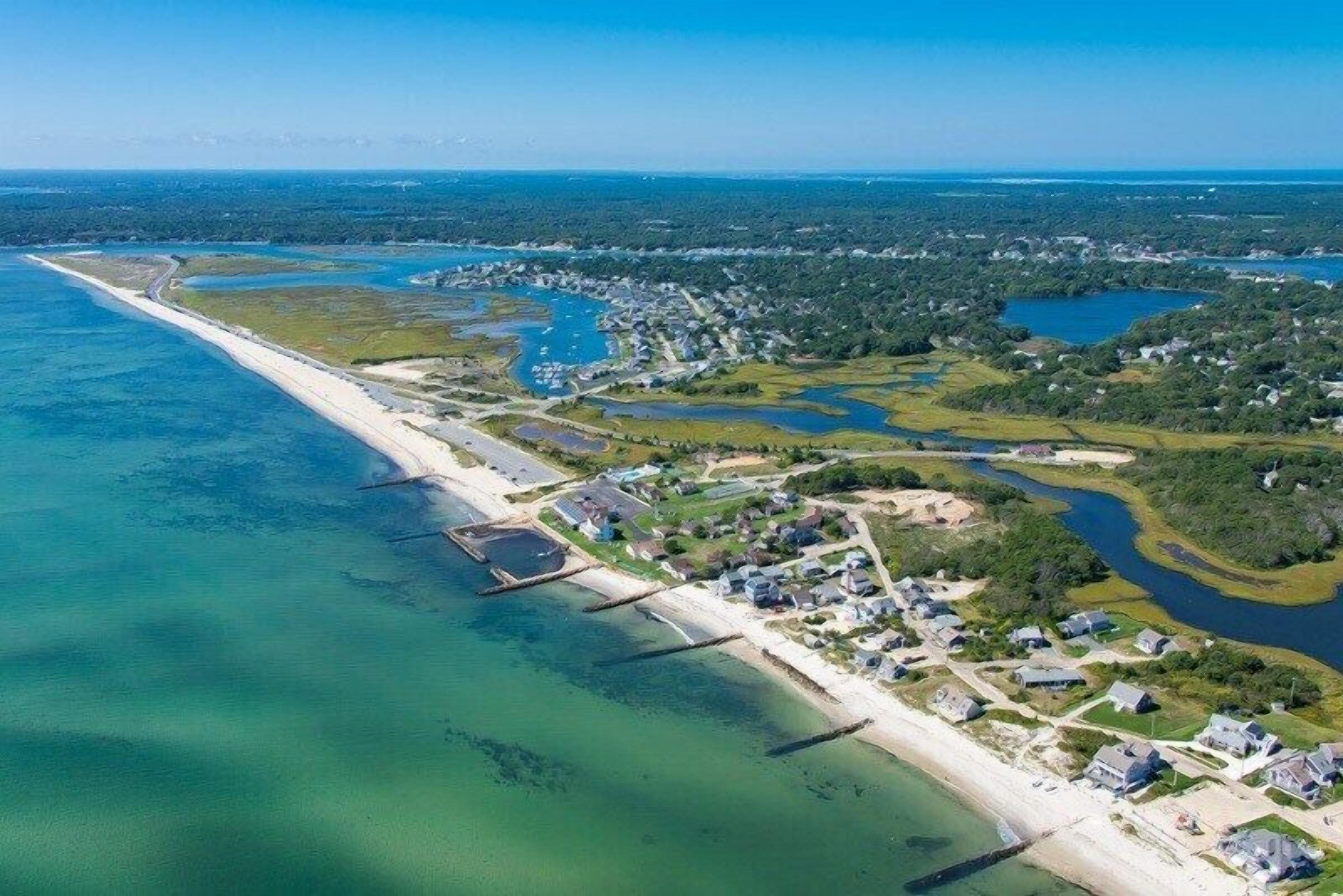 An aerial view of a coastal area with a long sandy beach, clear blue waters, and a mix of residential buildings and open green spaces.