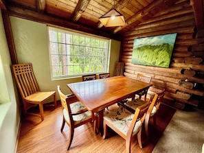 A wooden dining room with a large table and chairs, a window with a view of greenery, and a painting on the wall.