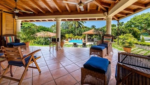 An outdoor patio with wicker furniture, a ceiling fan, and a view of a swimming pool and lush greenery.