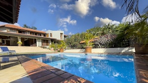 A swimming pool with a tiled deck, surrounded by potted plants and a building with a tiled roof.