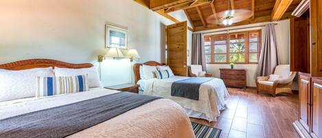 A hotel room with two beds, wooden ceiling beams, a window with curtains, a wicker chair, and a nightstand with lamps.