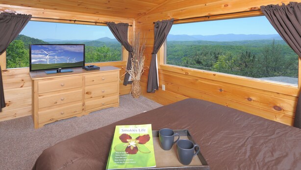 Une chambre dans un chalet en bois, avec un lit, une commode, une télévision et une fenêtre donnant sur un paysage de montagnes et d’arbres.