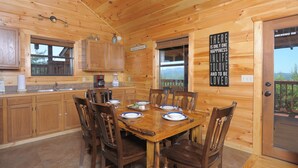 L’intérieur d’un chalet en bois comprend une table à manger pour quatre personnes, des armoires en bois, un coin cuisine et une grande fenêtre.