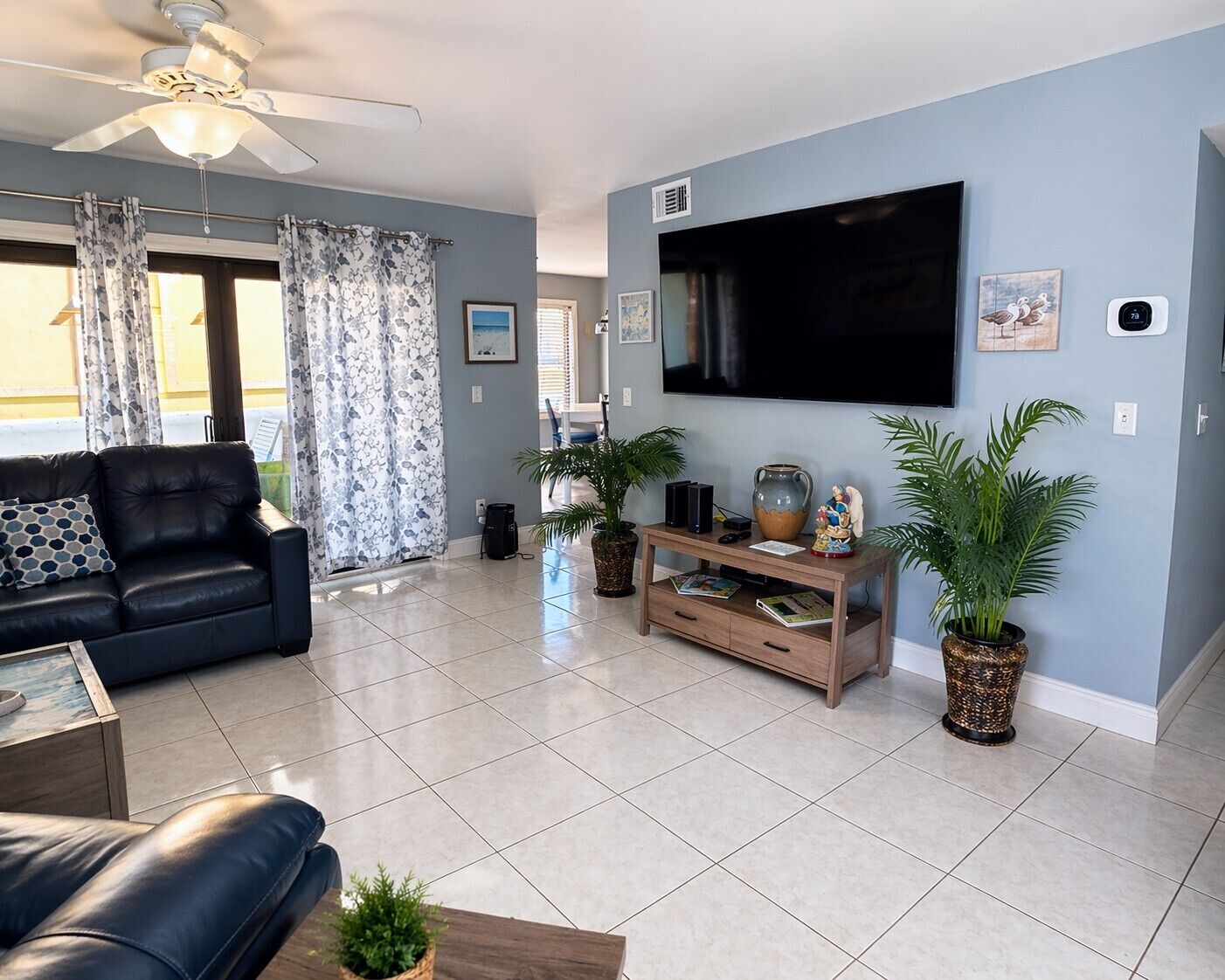 A living room with a black leather sofa, a wooden TV stand with a flat-screen TV, and two potted plants.