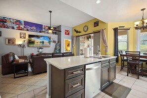 A modern kitchen with a central island, a dining area with a table and chairs, and a living room with a sofa and a TV wall.