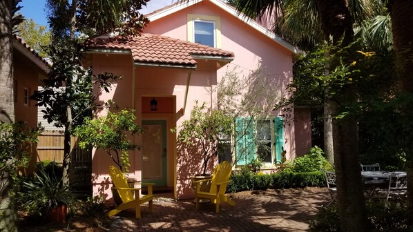 Une maison rose avec un toit en tuiles, deux chaises jaunes et un petit jardin agrémenté de différentes plantes.