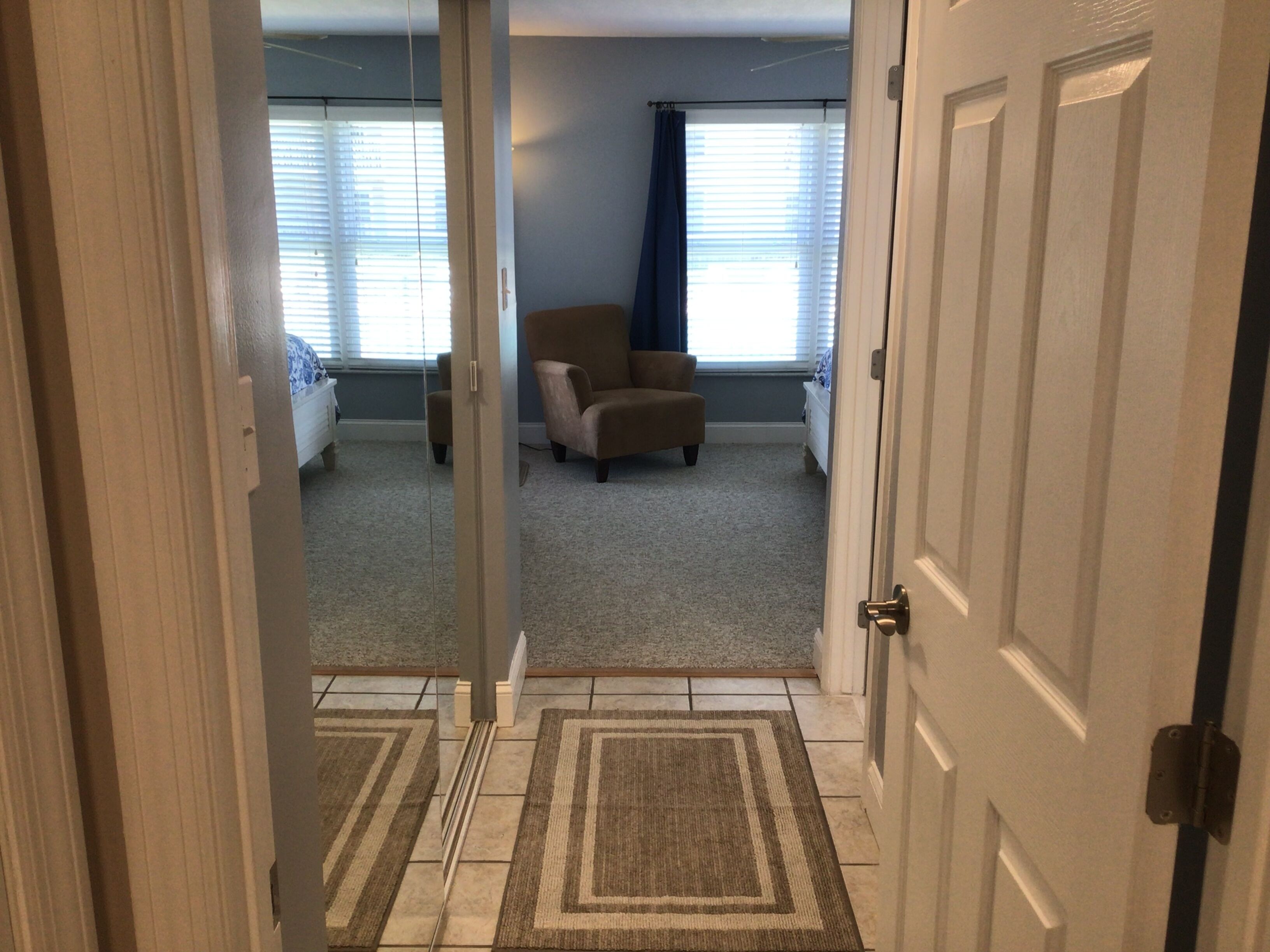 A hallway leading to a living room with a beige carpet, a brown and beige rug, a brown chair, and a window with blinds.