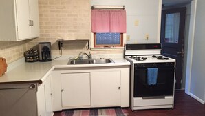 A compact kitchen with white cabinets, a stainless steel sink, a gas stove, and a window with a red and white valance.
