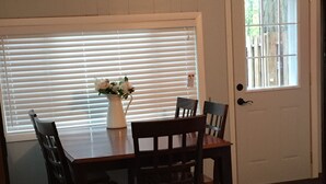 A dining area with a wooden table and chairs, a vase with flowers, and a window with blinds.