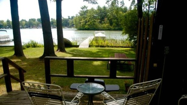 A deck with two metal chairs and a small table, overlooking a lake with a dock and a grassy area.