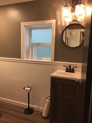 Un baño con ventana, lavamanos, espejo y dispensador de papel higiénico.