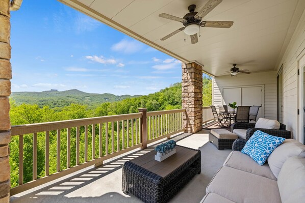 A covered porch with a stone pillar, wicker furniture, and a view of green hills.