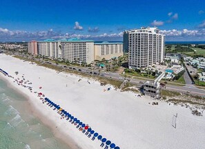 A coastal hotel with multiple high-rise buildings, a beach with umbrellas, and a clear view of the ocean.