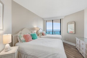 A bedroom with two beds, a bedside table with a lamp, a dresser, and a large window with a view of the ocean.