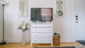 A white dresser with a flat-screen TV, two wall mirrors, a white vase with flowers, a floor lamp, and a wicker basket. - Quaint Downtown Condo (Salt Lake City)