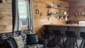 A cozy cabin interior with a wooden ceiling, a black leather sofa with patterned cushions, a wooden coffee table, and a kitchen area with a bar stool and shelves. - 3 Bed Cozy Cabin Near Trails, Tippy Dam, Manistee River, Casino. (Wellston)