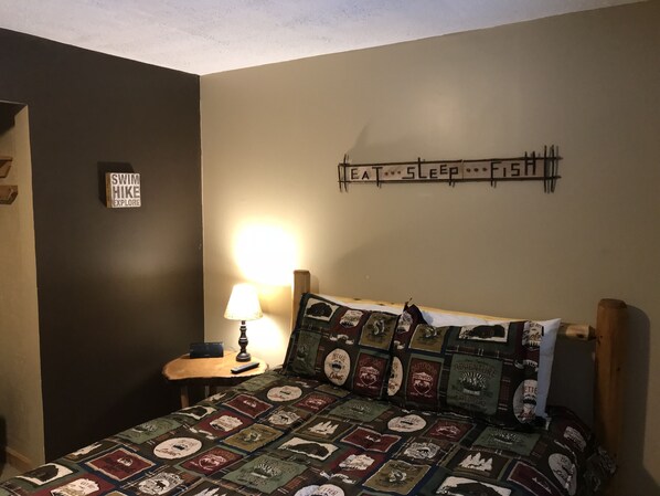 Un dormitorio con una colcha acolchada con parches de temática viajera, un cartel en la pared con la inscripción 'COMER, DORMIR, PESCAR', una lámpara sobre una mesita de noche y un cartel en la pared que dice 'NADAR, CAMINATAS, EXPLORAR'.
