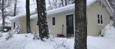 Una casa cubierta de nieve con puerta, ventanas y un cobertizo pequeño en un paisaje nevado.