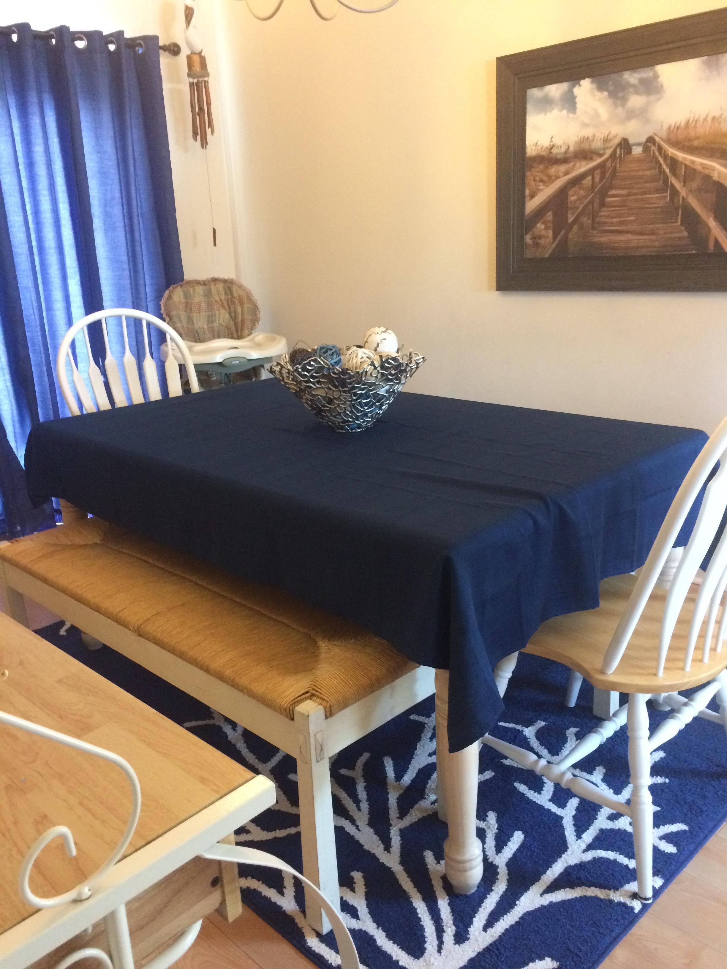 A dining area with a table covered by a dark cloth, surrounded by white chairs with wooden legs. A woven basket with decorative items is placed on the table.