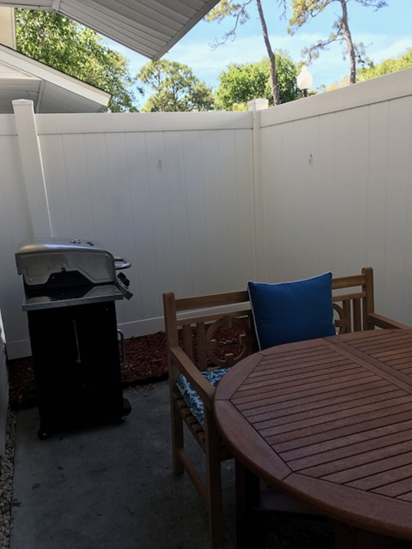 A backyard with a round wooden table, a barbecue grill, and a wooden bench with a blue cushion.