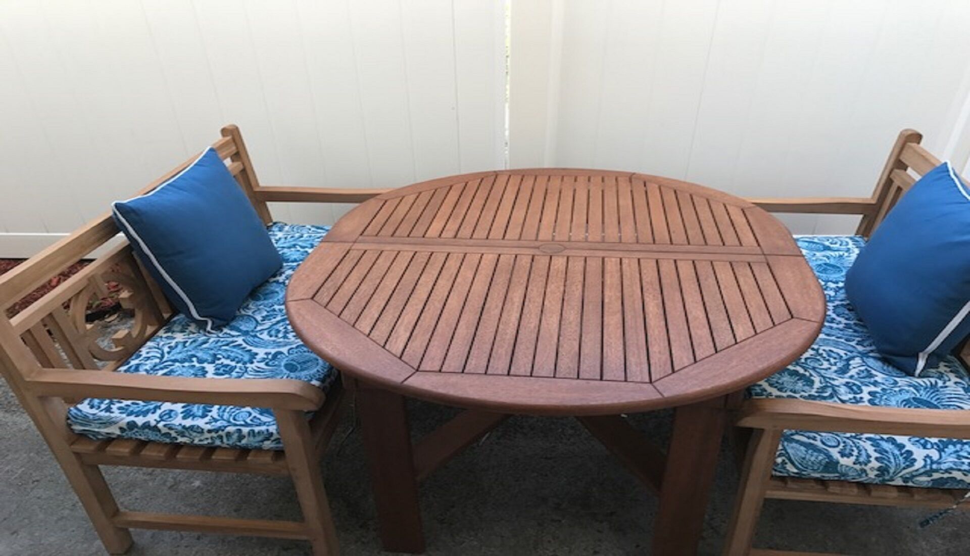 A round wooden table with a slatted top, flanked by two wooden chairs with blue patterned cushions.