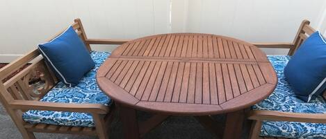 A round wooden table with a slatted top, flanked by two wooden chairs with blue patterned cushions.